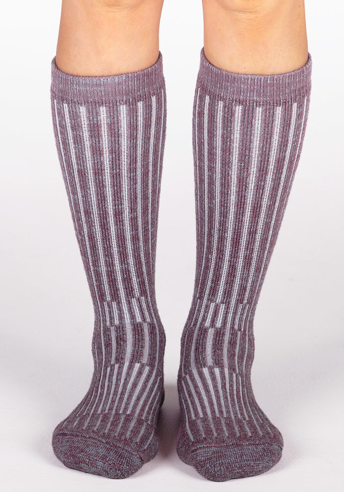 A pair of legs wears Woolx Womens Joey Mid Calf Socks in Wildflower, featuring cozy purple and gray vertical stripes, standing on a white surface.