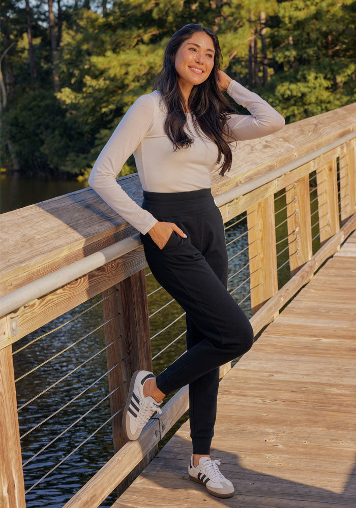 A woman with long dark hair wears a light long-sleeve top, Woolx Willa Joggers in black, and white sneakers as she smiles, leaning on a wooden bridge railing by the water with trees in the background.