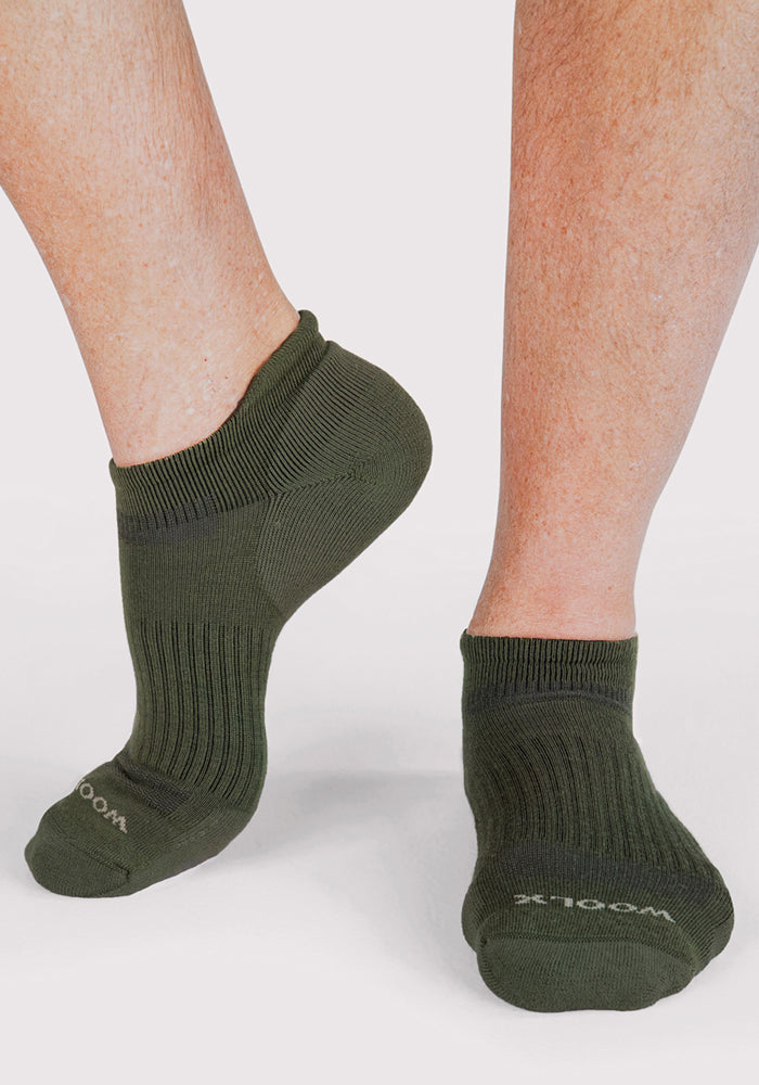 A close-up of two feet in Woolx Mens Rowan No Show Socks in Winter Moss, with one heel raised to show the soft Merino wool texture and snug fit. The background is plain and light-colored.
