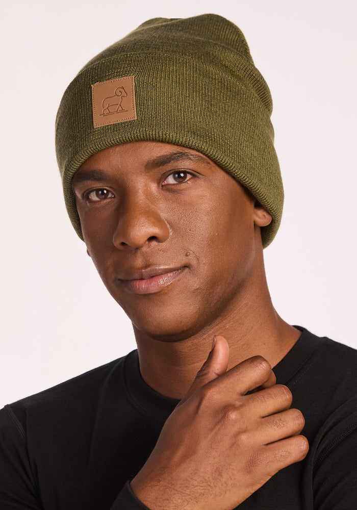 A person wearing the Woolx Baylor Beanie in Winter Moss, featuring a small tan patch, and a black shirt, looks at the camera with a neutral expression while touching the side of their face.