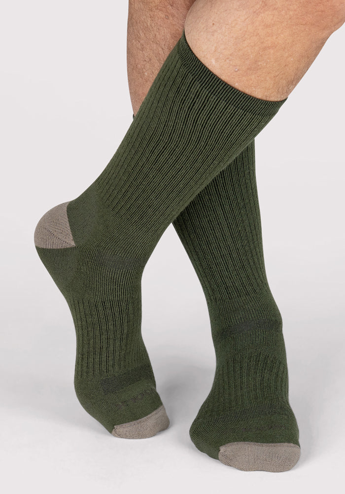 A person wearing Woolx Mens Core Crew Socks in Winter Moss, featuring ribbed olive green merino wool with light brown heels and toes, stands with one foot crossed over the other against a light background.