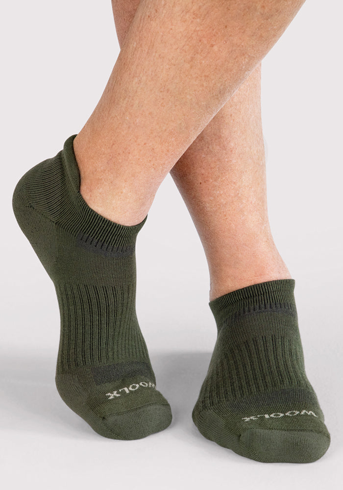 A person with crossed legs wears Woolx Mens Rowan No Show Socks in Winter Moss, standing against a light gray background. "WOOL" is visible on the toe of one cushioned, moisture-wicking Merino wool sock.