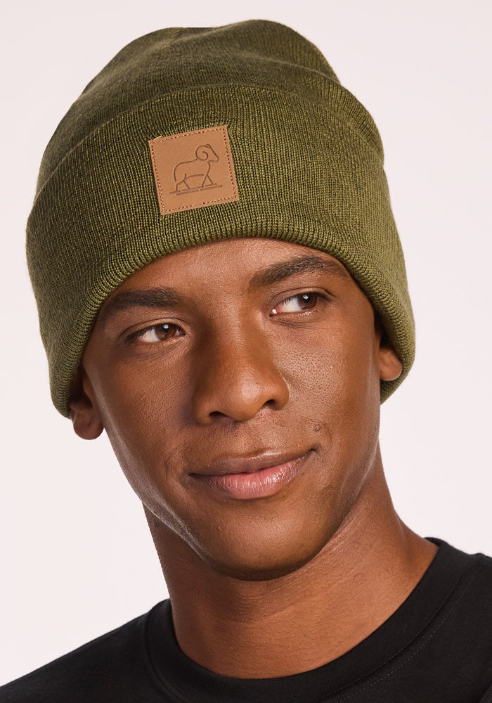 A person wearing the Woolx Baylor Beanie in Winter Moss, featuring a tan patch with a simple animal outline, paired with a black shirt and looking slightly to the side with a neutral expression.