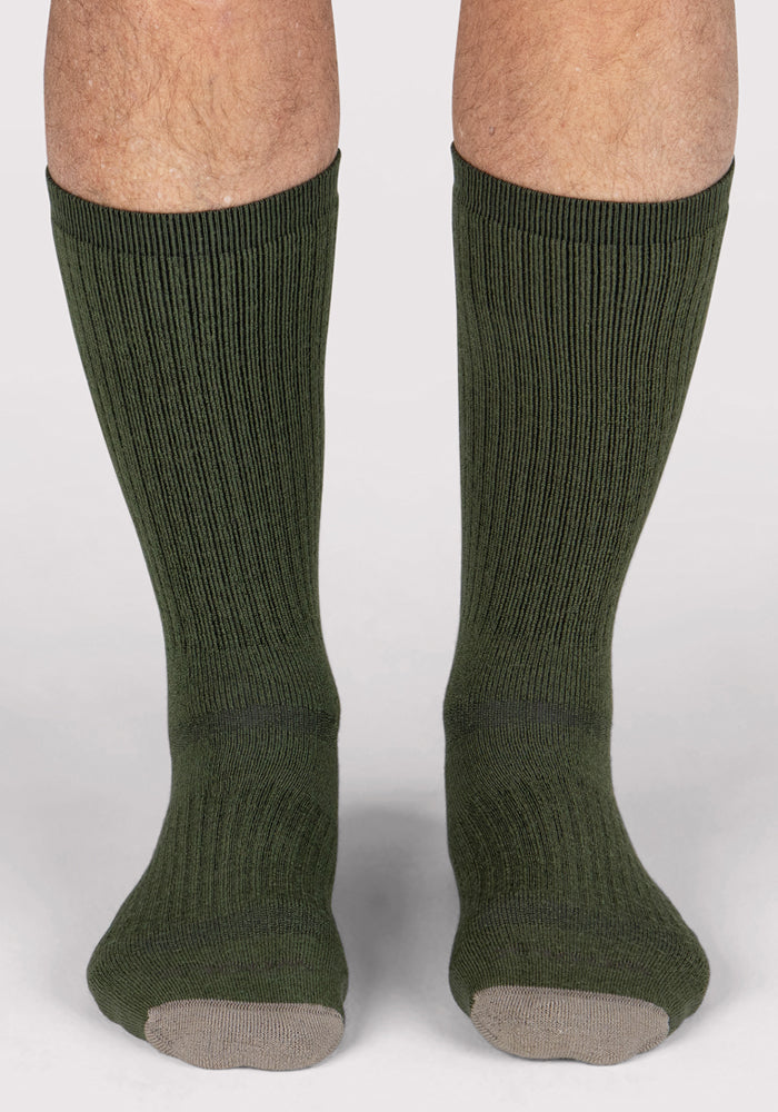 A pair of legs wearing Woolx Mens Core Crew Socks in Winter Moss, featuring moisture-wicking ribbed fabric and light gray toes, standing on a white surface.
