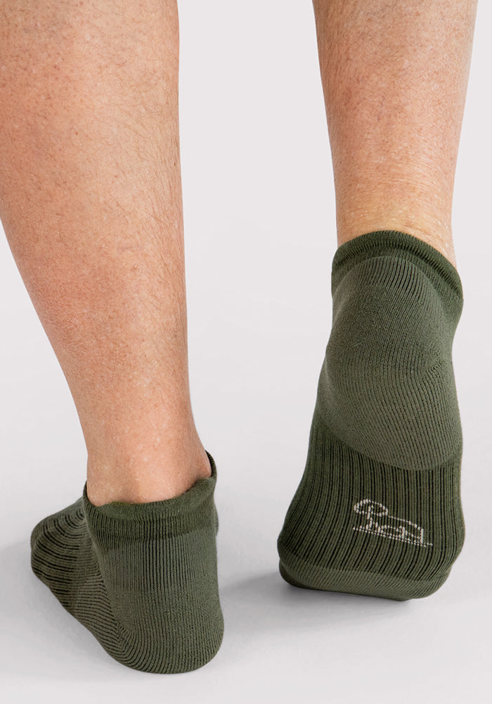 A close-up of a person's lower legs wearing Woolx Mens Rowan No Show Socks in Winter Moss. The olive green, moisture-wicking ankle socks feature cushioning and a small white logo on the heel. The background is light gray.