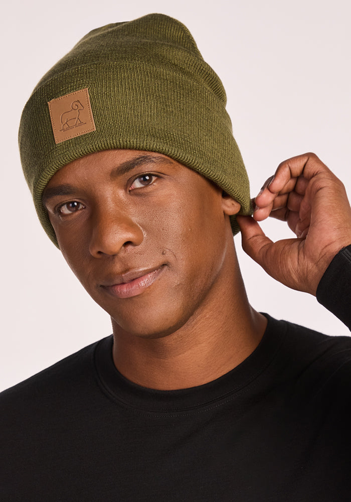 A person wearing the Woolx Baylor Beanie in Winter Moss with a small brown patch and a black shirt, looking at the camera and adjusting the beanie against a plain background.