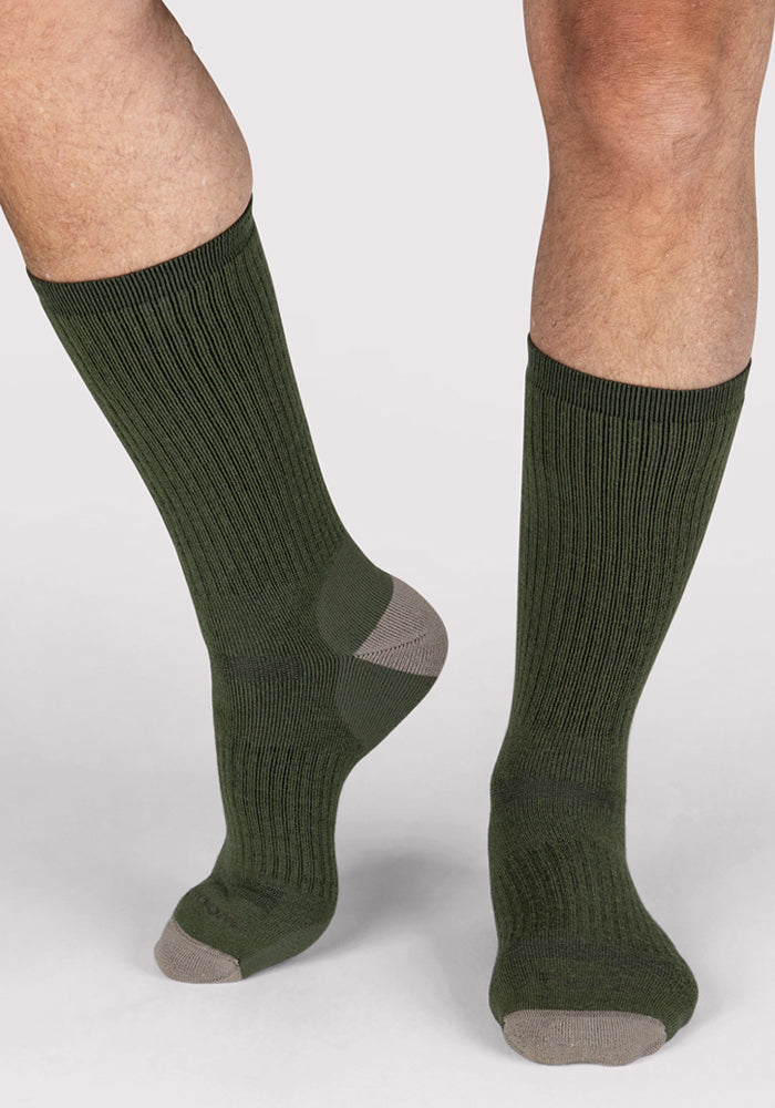 A person wears Woolx Mens Core Crew Socks in Winter Moss, featuring dark green merino wool with gray toe and heel accents. The odor-resistant, moisture-wicking crew socks cover the lower legs and feet against a light background.