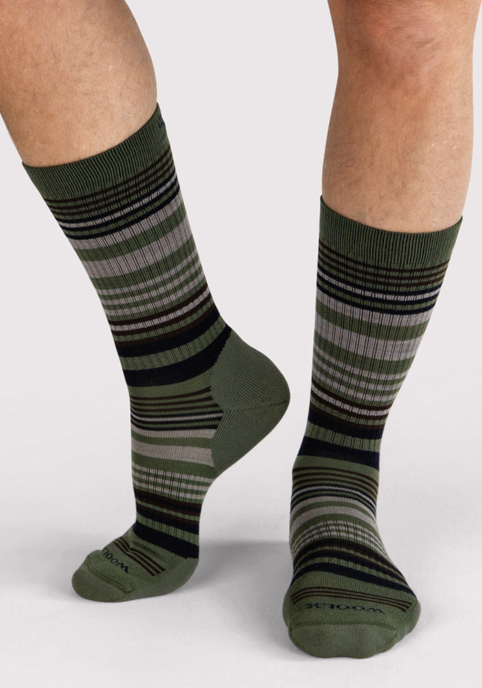 A person models the Woolx Mens Everyday Crew Socks in Winter Moss Stripe, featuring green, black, and gray horizontal stripes. The moisture-wicking socks are shown mid-calf on the lower legs and feet against a light background.
