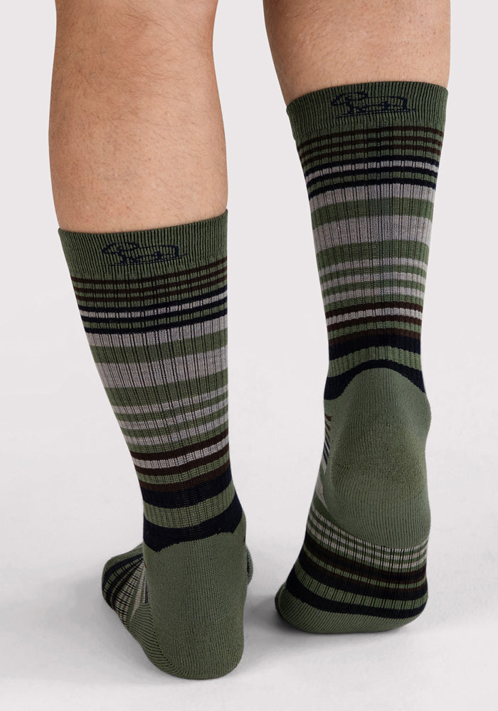 A person stands on a light surface, showing the back of their lower legs and feet while wearing Woolx Mens Everyday Crew Socks in Winter Moss Stripe—green cushioned crew socks with black, gray, and white horizontal stripes.