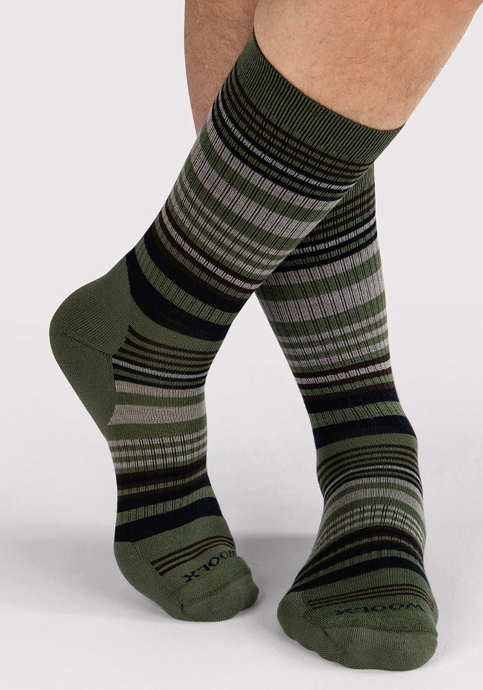 A person wearing Woolx Mens Everyday Crew Socks in Winter Moss Stripe, featuring black, gray, and beige stripes, stands with one leg crossed over the other against a light gray background.