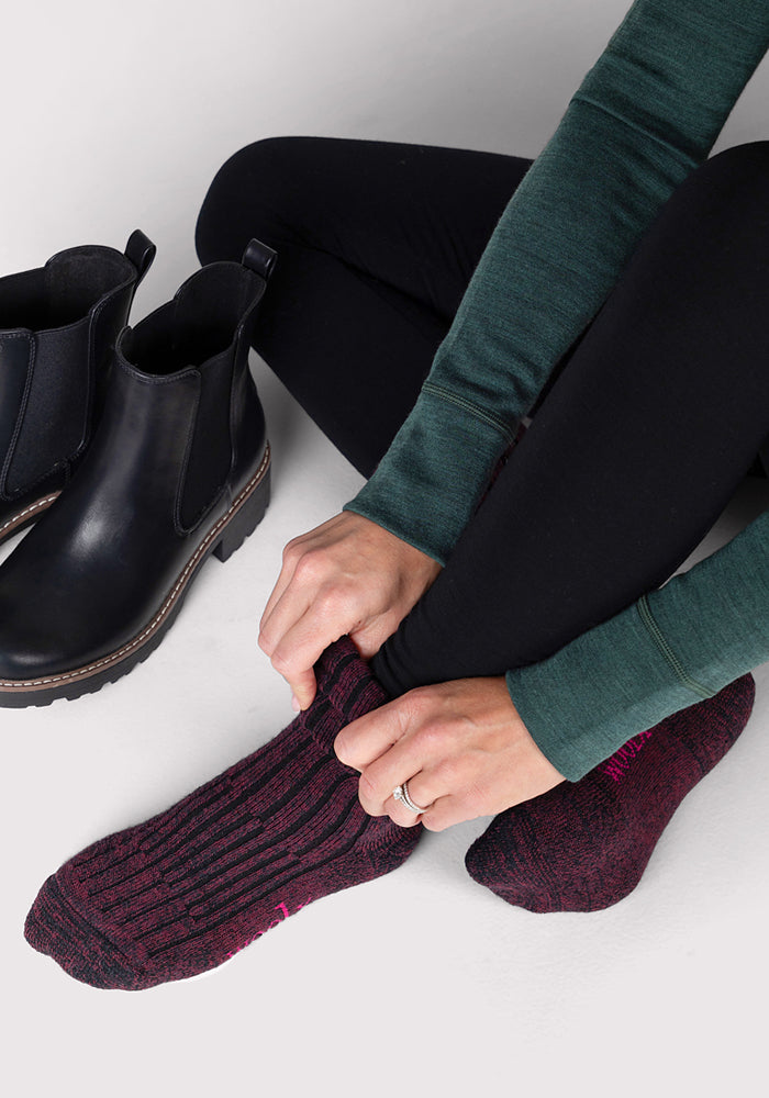 Wearing black leggings and a green long-sleeve top, a person puts on Woolx Womens Joey Crew Socks in Warm Violet, sitting beside black ankle boots on a light floor.
