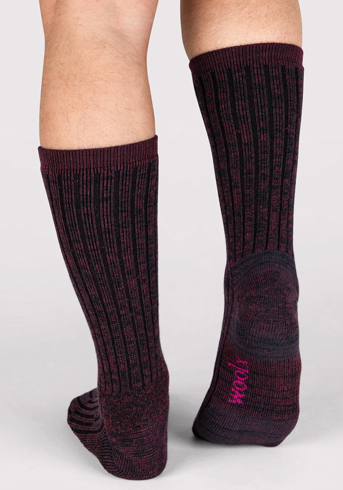 A person wearing Woolx Mens Joey Crew Socks - Warm Violet stands on a light surface. These cozy socks have a ribbed texture and pink text on the sole. Only the lower legs and feet are visible.