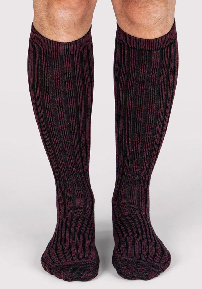 A pair of legs wearing Woolx Mens Joey Mid Calf Socks in Warm Violet, featuring a moisture-wicking, dark purple and black vertical striped pattern, standing on a light-colored surface.