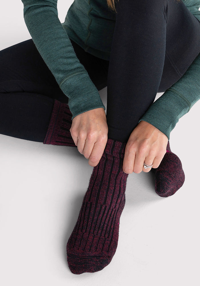 A woman in black leggings and a green long-sleeve top sits on the floor, pulling on Woolx Womens Joey Crew Socks in Warm Violet. Only her lower body and hands are visible.