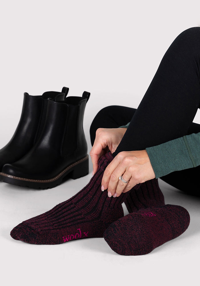 A person sits on the floor pulling on Woolx Womens Joey Crew Socks in Warm Violet, featuring "wool" in pink on the sole. Black ankle boots sit nearby, and the person wears black leggings and a green long-sleeve top.