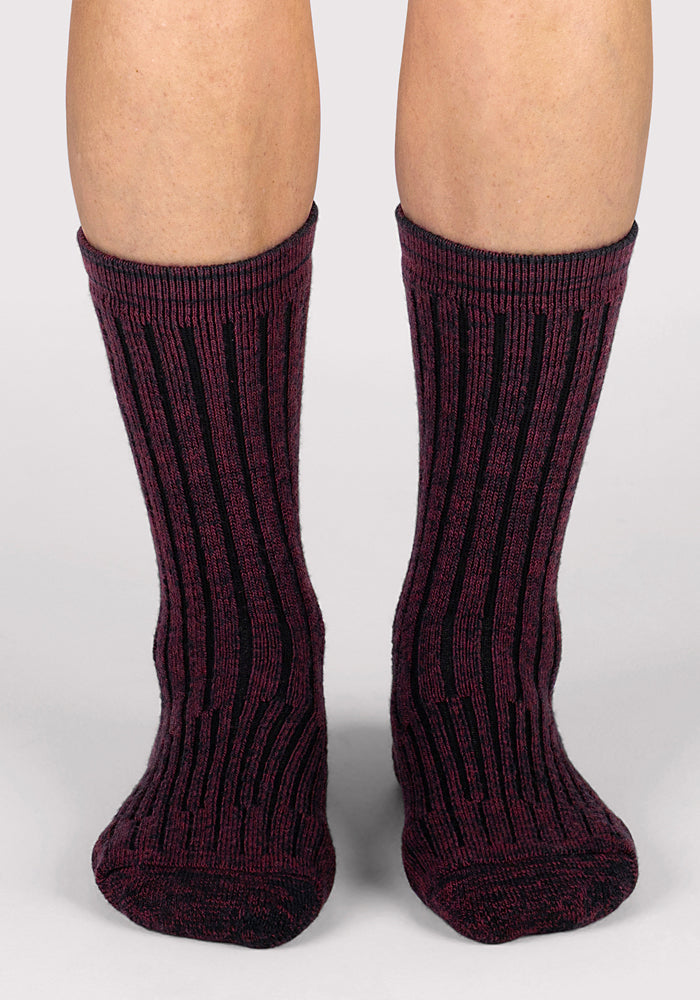A pair of feet wearing Woolx Womens Joey Crew Socks in Warm Violet, featuring a maroon and black ribbed design, stand on a light-colored surface with toes pointed forward.