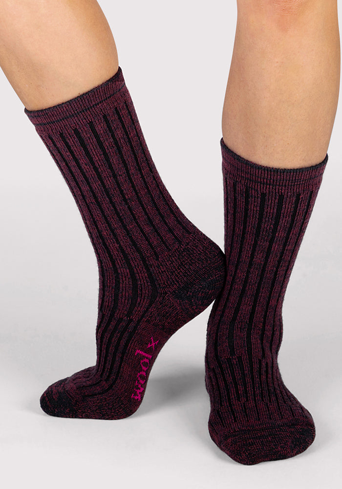 A person wears Woolx Womens Joey Crew Socks in Warm Violet, featuring ribbed texture, black vertical stripes, and a pink logo on the sole. The background is light gray.