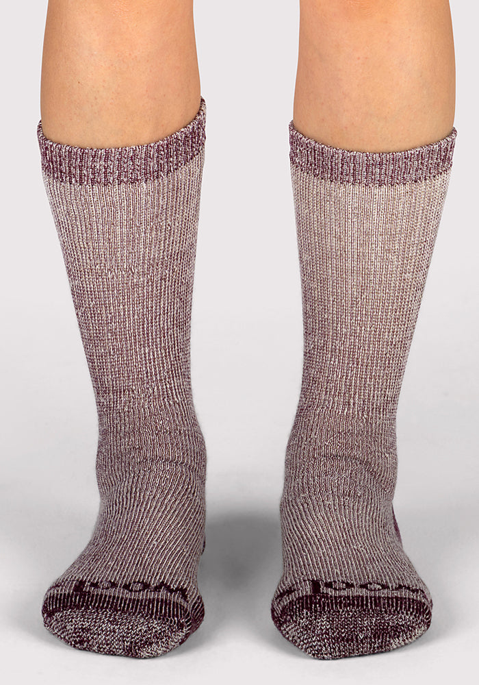 A close-up of a person's lower legs showcases Woolx Womens Extreme Crew Socks in Windsor Wine, featuring thick, marled purple and gray knit, worn on a light gray surface.