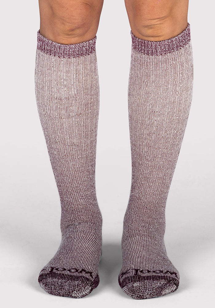 A person wearing Woolx Womens Extreme Over The Calf Socks in Windsor Wine stands on a plain, light-colored surface, with only their legs and feet visible.