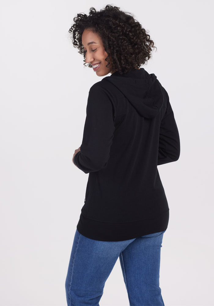 A person with curly hair is wearing a black Ryann Hoodie by Woolx and blue jeans. They are smiling and looking over their shoulder against a plain, light background.