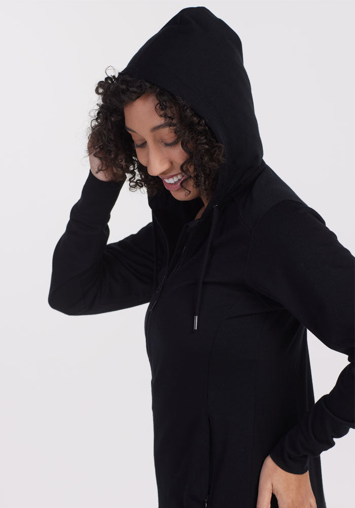 A person with curly hair is wearing a Woolx Ryann Hoodie in a lightweight, athletic fit, smiling and looking down slightly. The light gray background creates a soft contrast with the dark clothing.