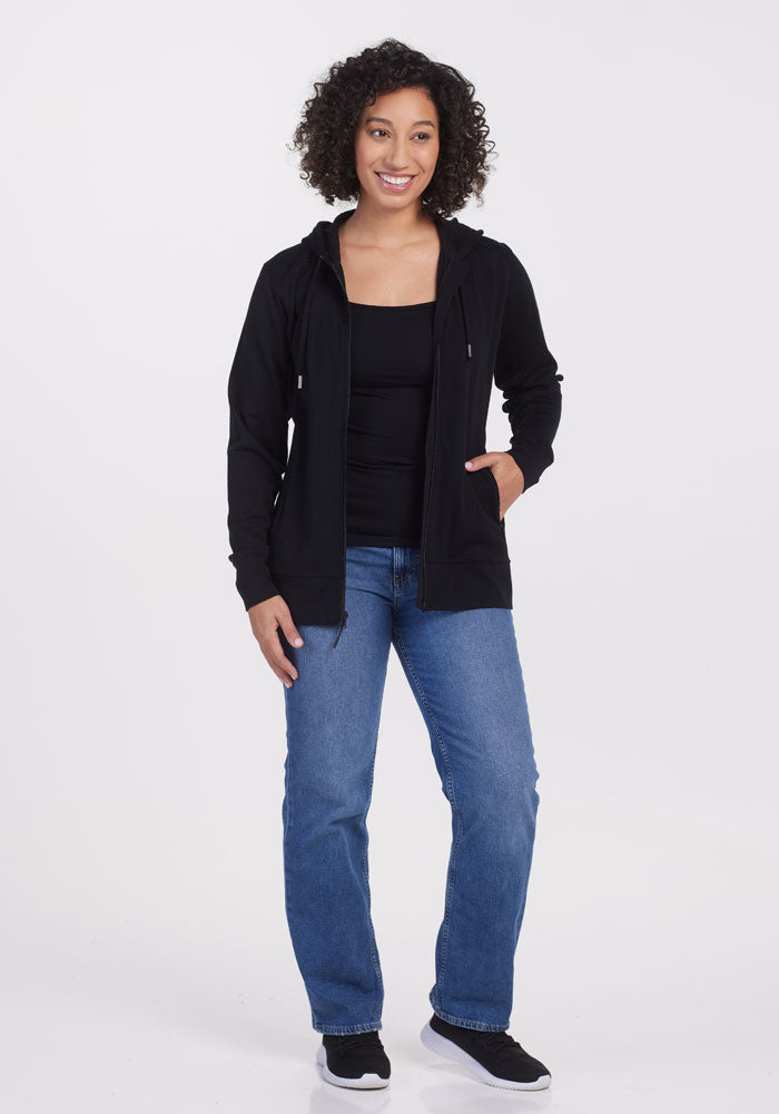 A person with curly hair smiles while standing in a sleek, athletic fit Ryann Hoodie OLD by Woolx. They pair it with a black top, blue jeans, and black sneakers. The backdrop is a crisp, plain white.
