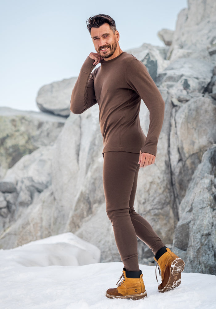 A man in Woolx Backcountry Leggings - Black stands on snow with rocky cliffs behind him, smiling and looking over his shoulder.