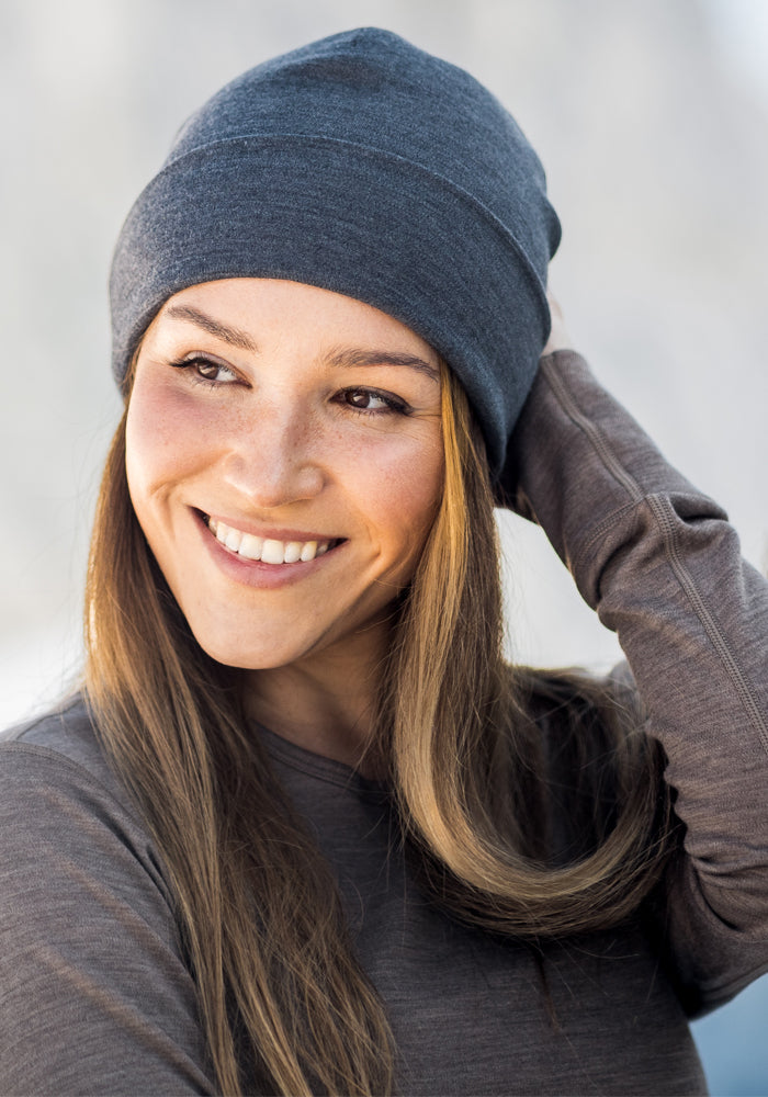 A woman with long brown hair smiles while wearing the Woolx Journey Hat in Steel Blue and a brown long-sleeve shirt, looking slightly to the side and touching her beanie. The softly blurred background adds warmth to the scene.