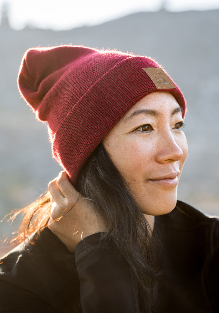 A woman in the Woolx Baylor Beanie - Rhubarb and a black jacket smiles softly while looking to the side outdoors against a blurred, sunlit background.