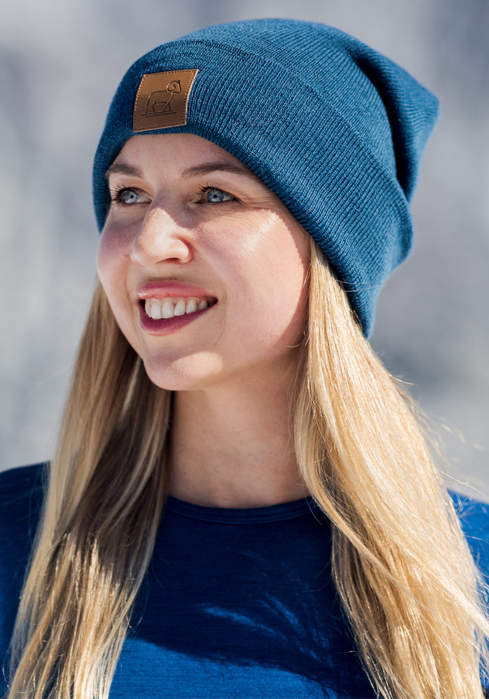 A woman with long blonde hair, wearing the Woolx Baylor Beanie in Majolica Blue and a matching shirt, smiles while looking to the side against a softly blurred, light-toned outdoor background.