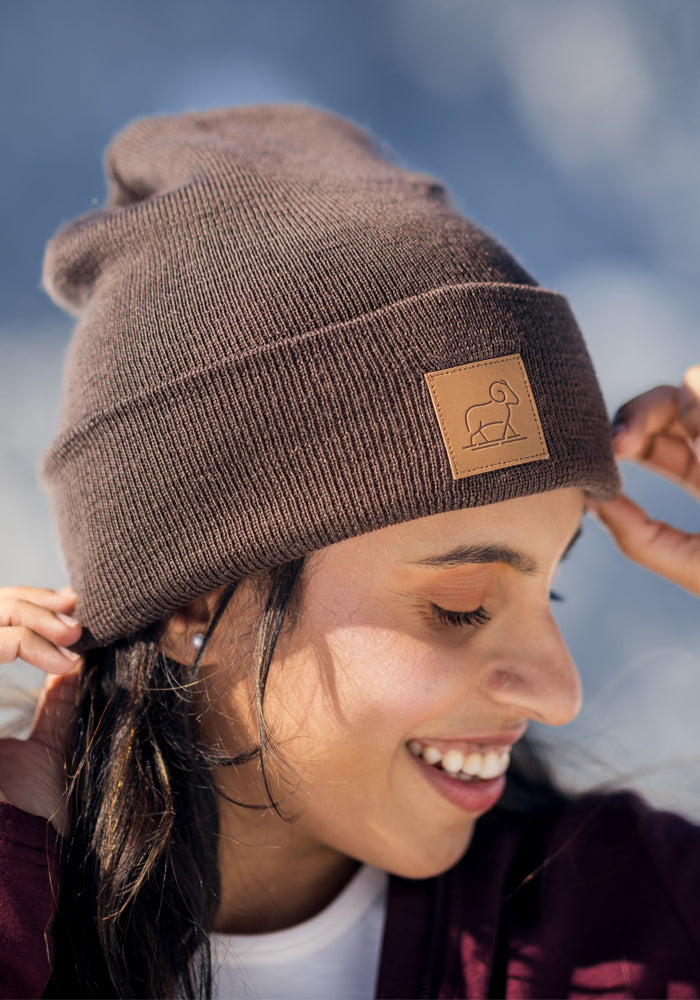 A smiling person adjusts the Woolx Baylor Beanie - Bracken, a brown merino wool winter hat with a square animal patch, while outdoors on a sunny day.