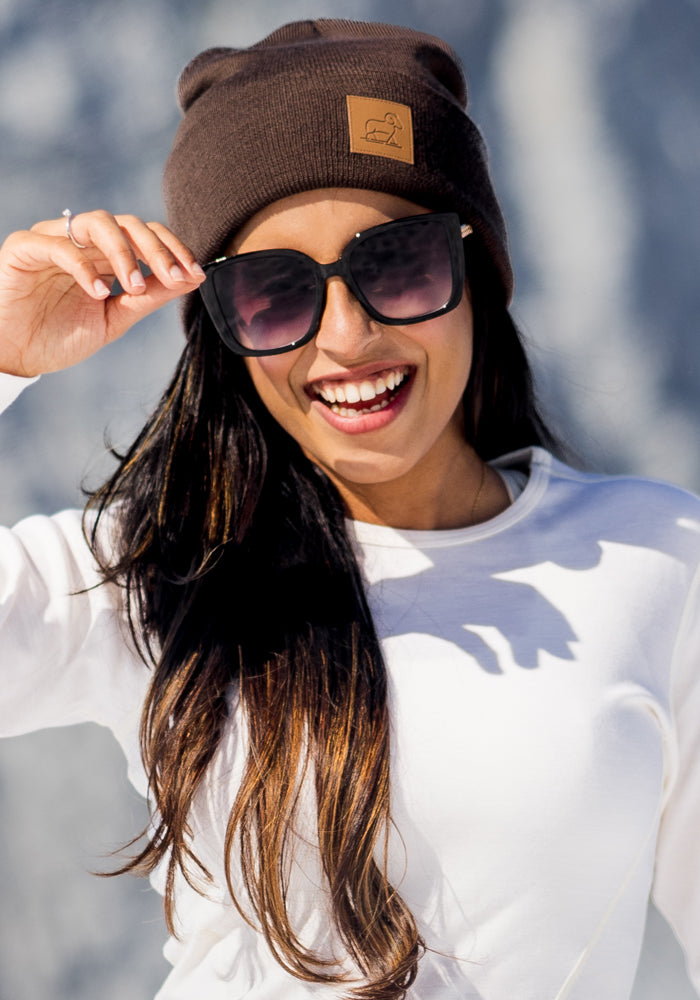 A woman with long dark hair smiles brightly outdoors, wearing the Woolx Baylor Beanie in Bracken, black sunglasses, and a white shirt against a blurred gray background as she holds her sunglasses.