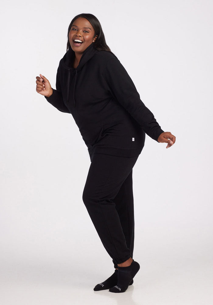 A woman cheerfully poses against a white background in Woolx Parker Sweatpants - Black, a black hoodie, and black socks, highlighting comfort and temperature regulation.