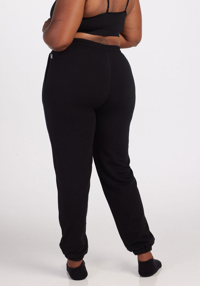 A person is barefoot, facing away from the camera against a white background, wearing Woolx Parker Sweatpants in black and a matching black sports bra.