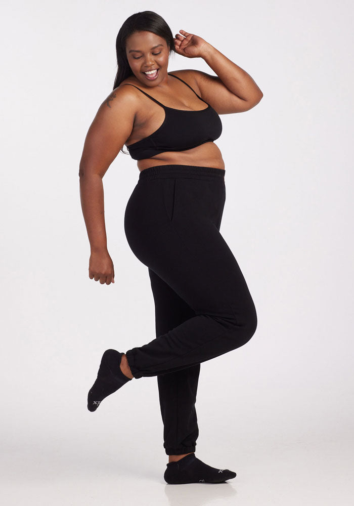A woman poses confidently against a white background, wearing a black sports bra, black socks, and Woolx Parker Sweatpants in black, made from Merino wool for optimal temperature regulation.