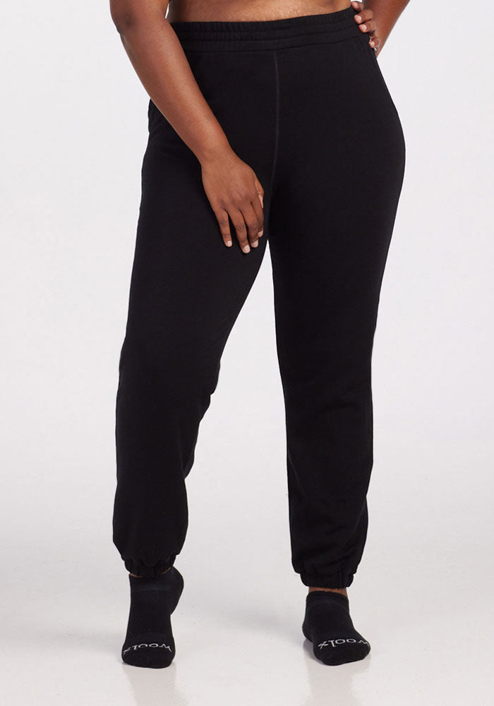 A person stands against a plain background wearing Woolx Parker Sweatpants in black and black socks, with their left hand resting on their right thigh. Only the lower half of their body is shown. 