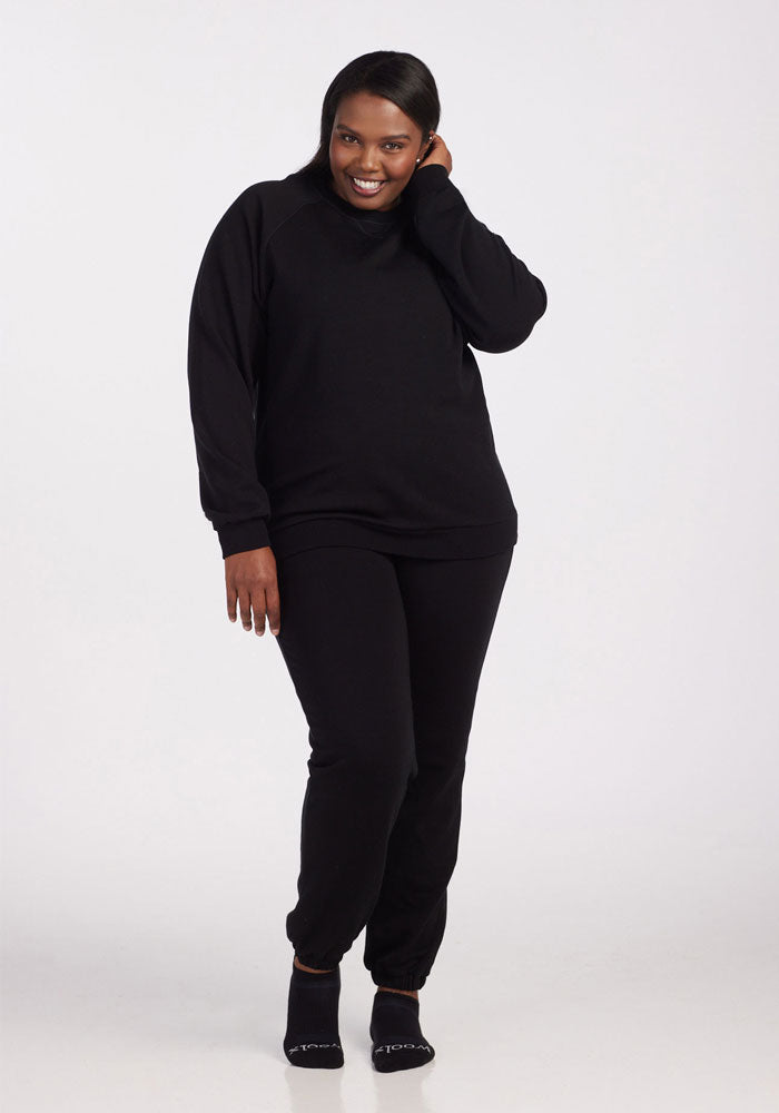 A woman smiles, touching her hair, dressed in a black long-sleeve top and Woolx Parker Sweatpants - Black (moisture-wicking Merino wool), with black socks, standing against a plain white background.