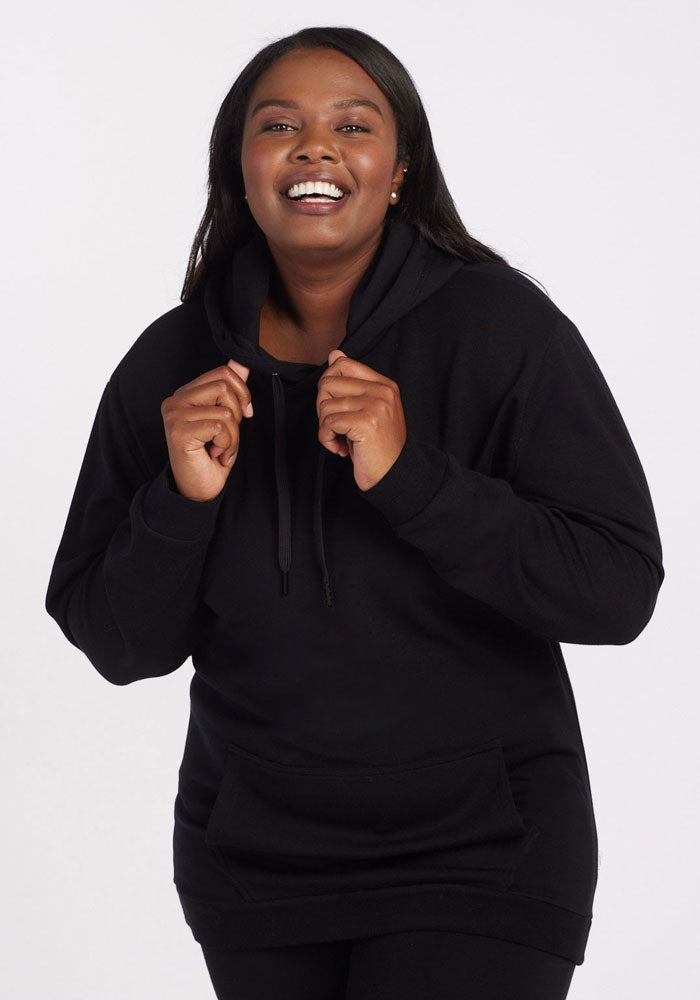 A woman smiles while holding the drawstrings of her black Woolx Avery Hoodie, standing against a plain white background.