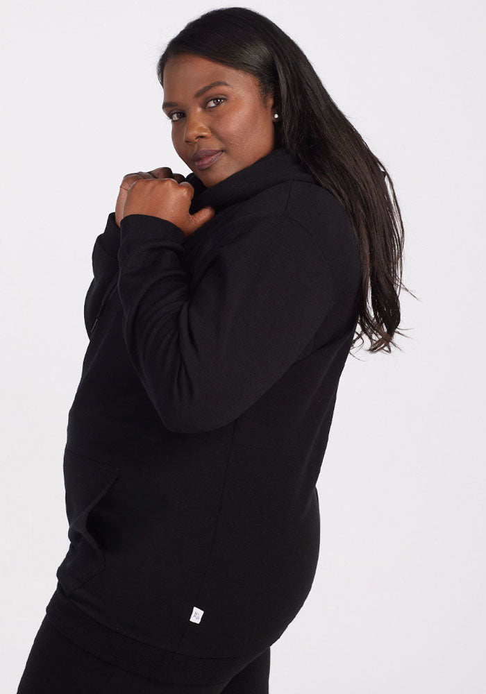 A woman with long dark hair models the Woolx Avery Hoodie in black, featuring a high collar, standing in side profile against a white background and smiling slightly at the camera.