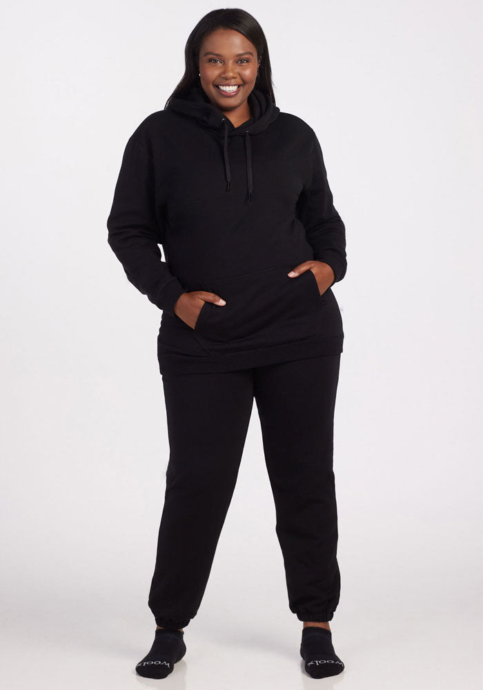 A smiling woman wears the Woolx Avery Hoodie in black, with hands in the front pocket, paired with black sweatpants and black socks, against a plain white background.