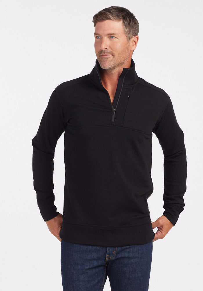 A man in a Woolx Mason 1/4 Zip - Black and blue jeans stands with hands on hips, looking to the side against a white background; the odor-resistant Merino wool fleece provides comfort and style. 