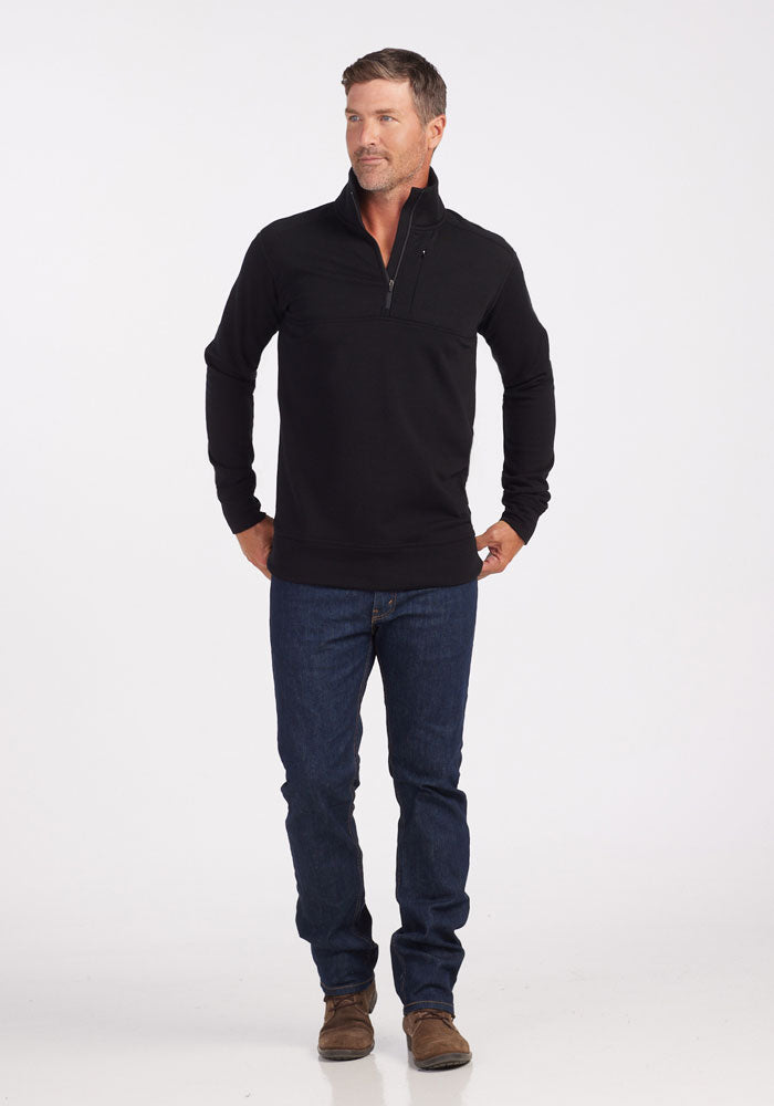 A man with short hair stands against a white background wearing the Woolx Mason 1/4 Zip - Black, made from odor-resistant Merino wool fleece, paired with dark blue jeans and brown shoes. He looks slightly left with his hands in his pockets.