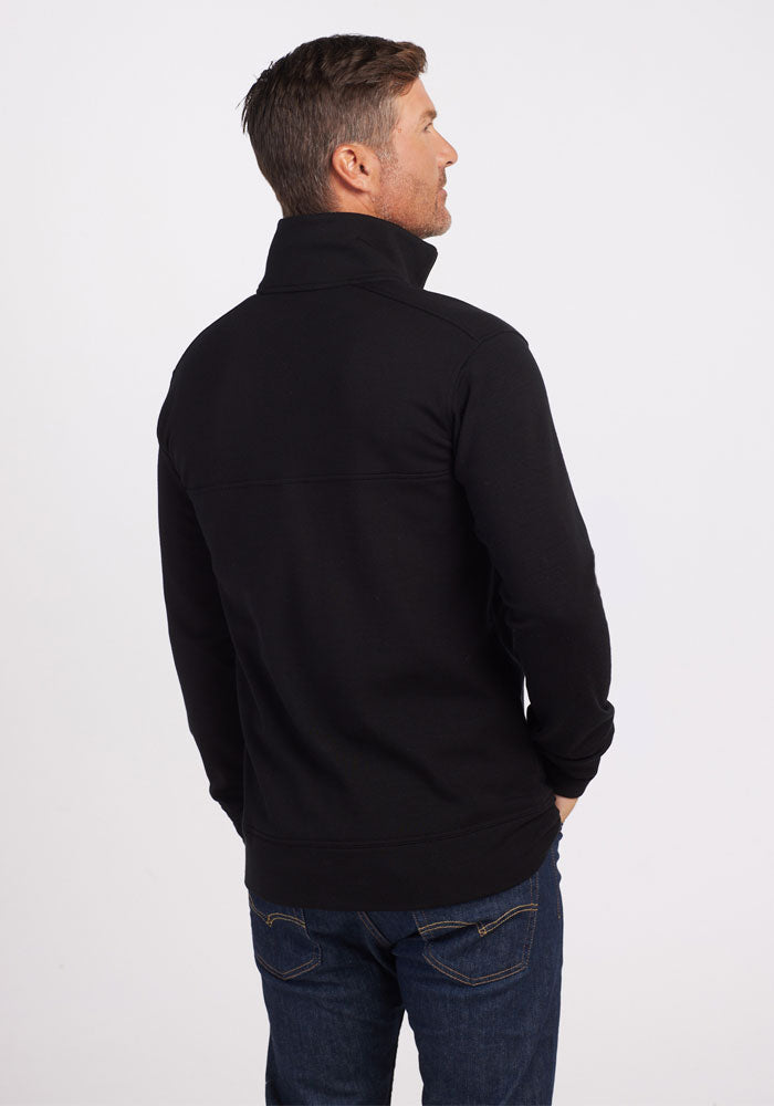 A man wearing a black Woolx Mason 1/4 Zip pullover and blue jeans stands with his back to the camera against a plain white background.
