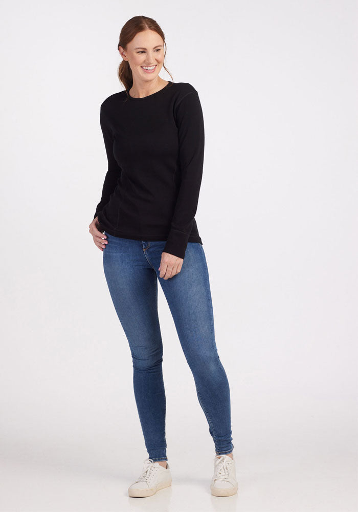 Smiling woman wears Woolx Hannah Petite Long Sleeve Top in black, blue jeans, and white shoes against a white background.