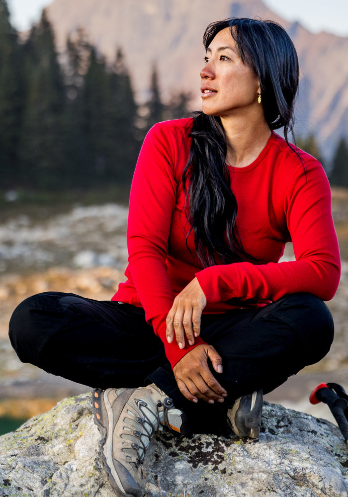 A woman wearing the Woolx Hannah Long Sleeve Top in Maraschino Cherry and hiking boots sits cross-legged on a rock outdoors, glancing to the side with trees and mountains blurred in the background.