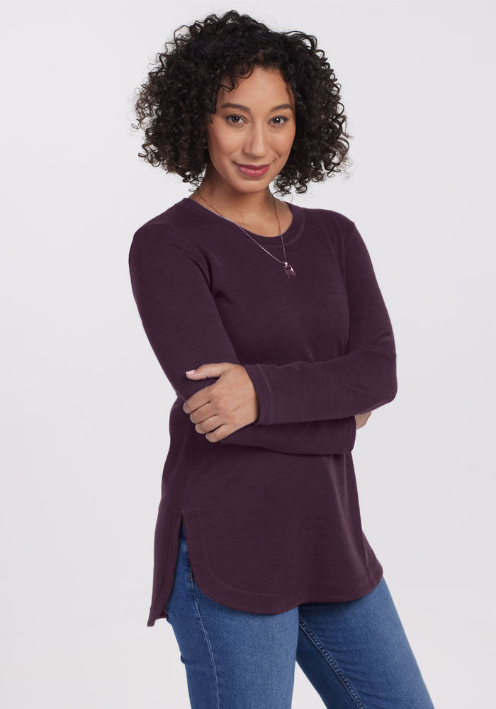 A curly-haired person smiles with arms crossed, showing off the relaxed fit of the Woolx Blair Tunic in Deep Plum, paired with blue jeans against a plain white background.
