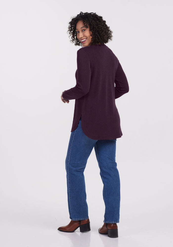 A curly-haired person smiles while turned slightly to the side, wearing a Woolx Blair Tunic in Deep Plum, blue jeans, and brown ankle boots against a light gray background.