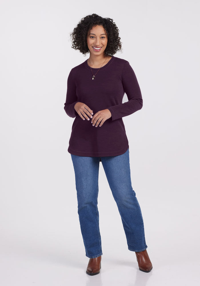 A woman with curly hair, wearing a Blair Tunic by Woolx in Deep Plum and blue jeans, smiles confidently. She's styled in a relaxed fit with brown ankle boots and a pendant necklace against a plain white background.