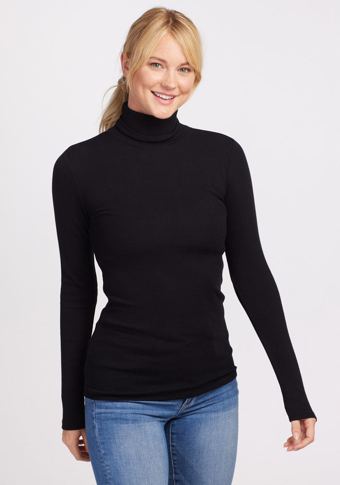Smiling woman in a Woolx Sage Petite Ribbed Turtleneck - Black and blue jeans stands against a white background.