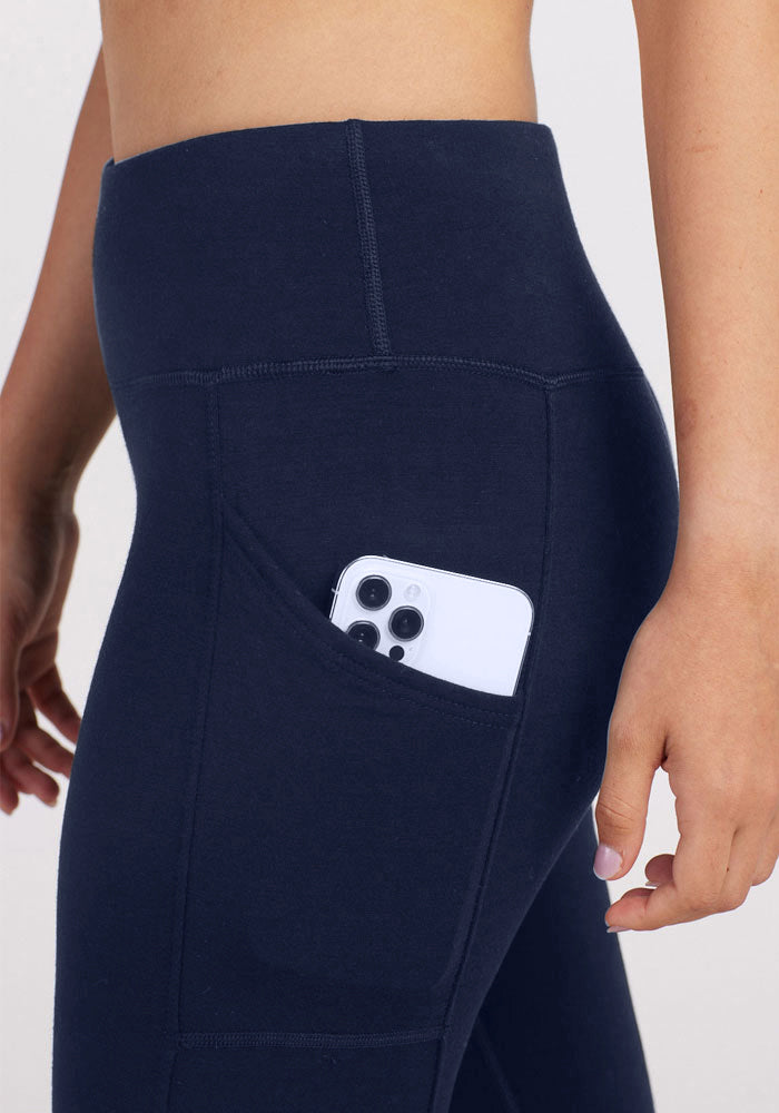 A person wears Woolx Piper Pocket Leggings in Deep Navy, with a smartphone partly tucked into the side pocket. Only their lower torso and one arm are shown.